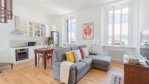 A modern living room with a grey sofa, wooden dining table, and a kitchen area with white cabinets and a microwave. - Rooftop Nest in Como City Center (Como)