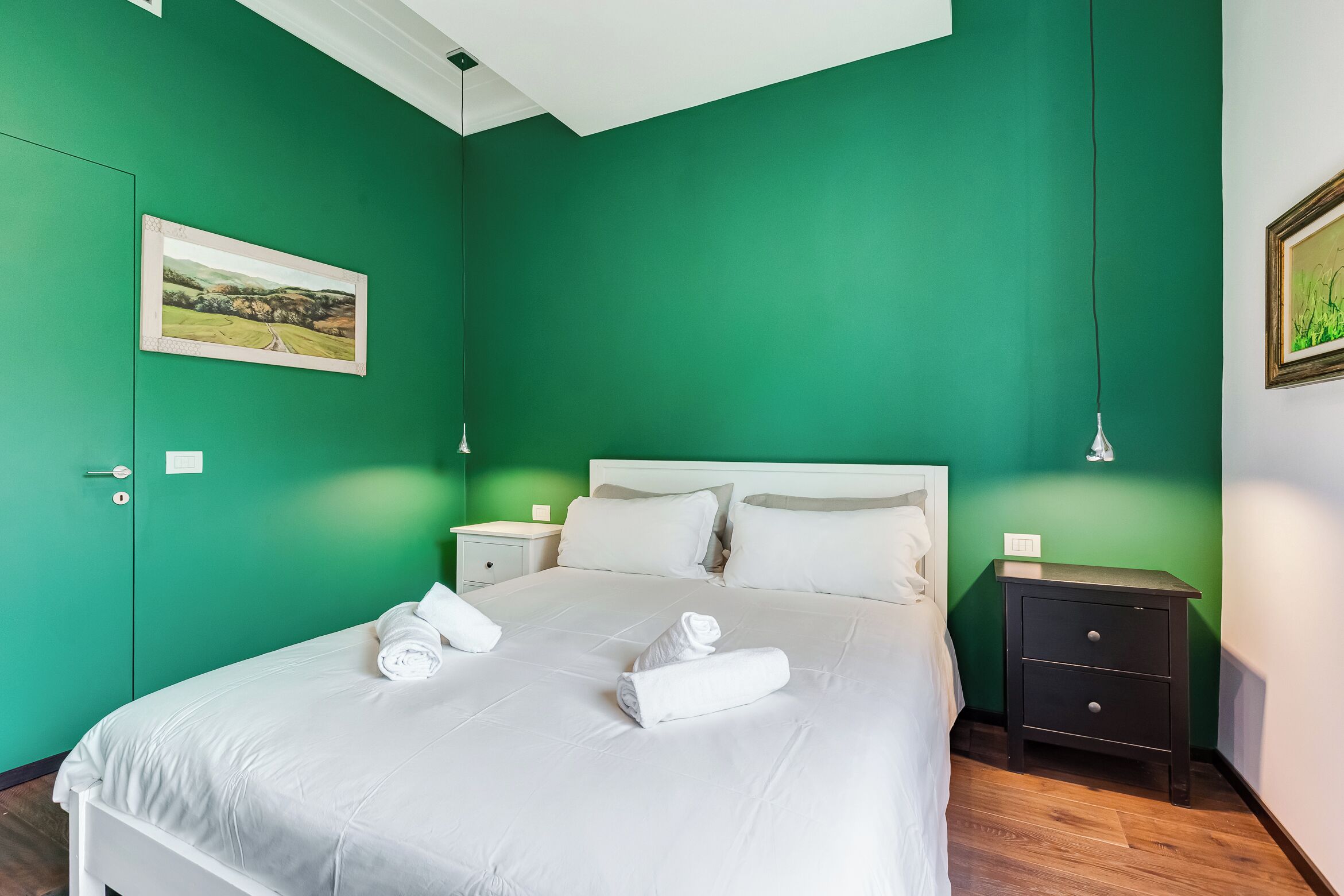 A neatly made bed with white linens and towels, a green wall, a small bedside table with a lamp, and a framed picture on the wall.
