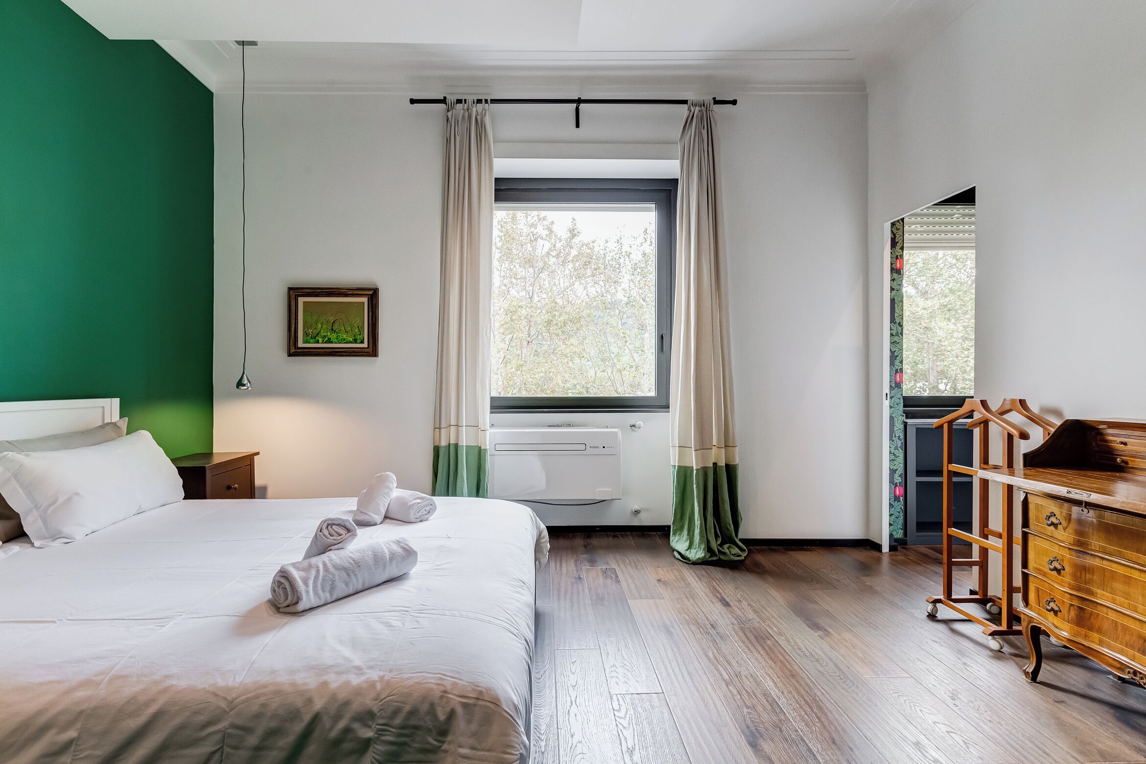 Una habitación de hotel con cama, cómoda, ventana con cortinas, radiador y un cuadro en la pared.