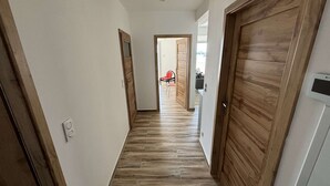 Ein Flur mit Holzdielen und Türen, der zu einem Raum mit einem roten Sessel und einem schwarzen Möbelstück führt.