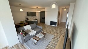 Ein modernes Wohnzimmer mit grauem Sofa, Couchtisch und Fernseher. Im Hintergrund ist eine Küchenzeile mit Spüle und Schränken zu sehen.