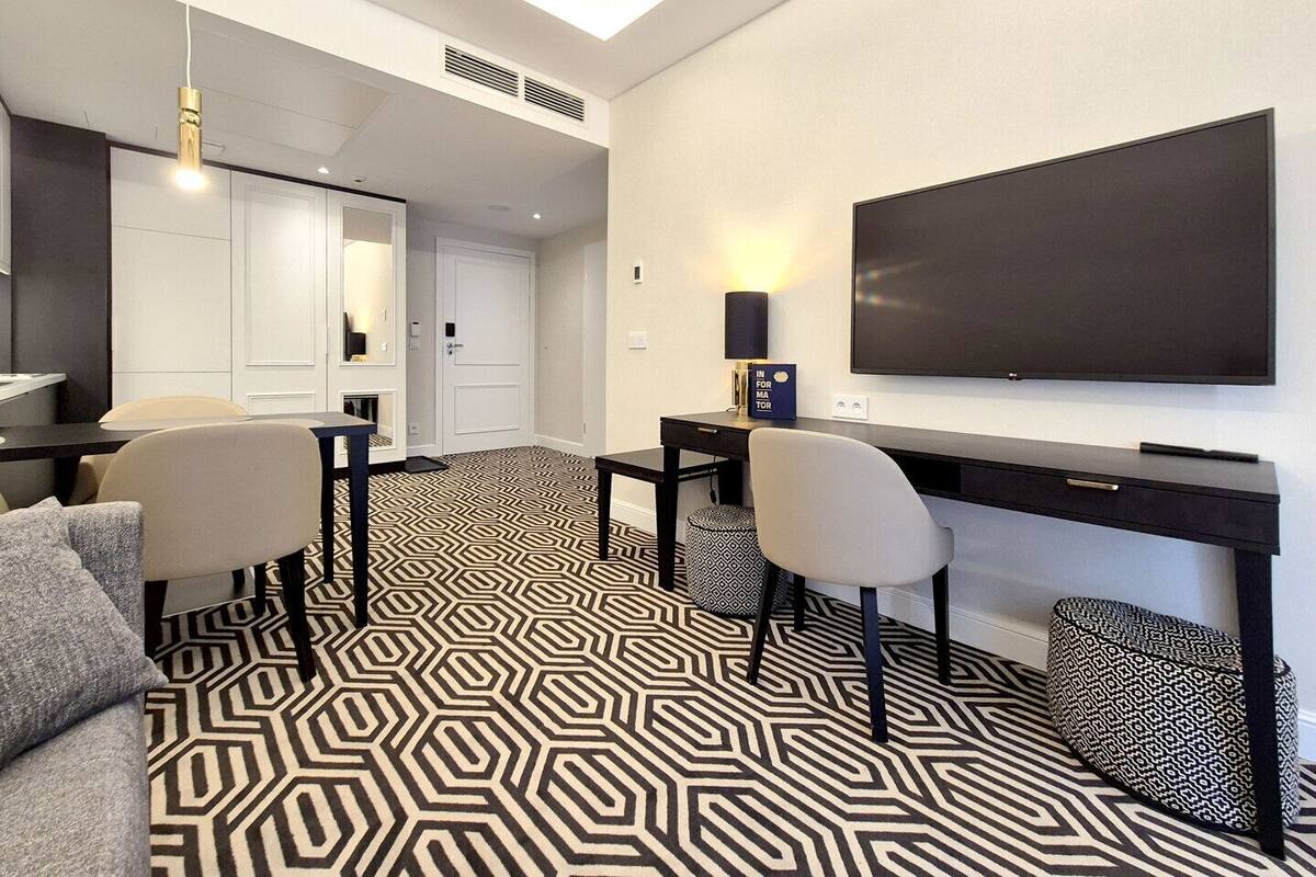 A modern hotel room with a large flat-screen TV mounted on the wall, a desk with a chair, a sofa, and a patterned carpet.