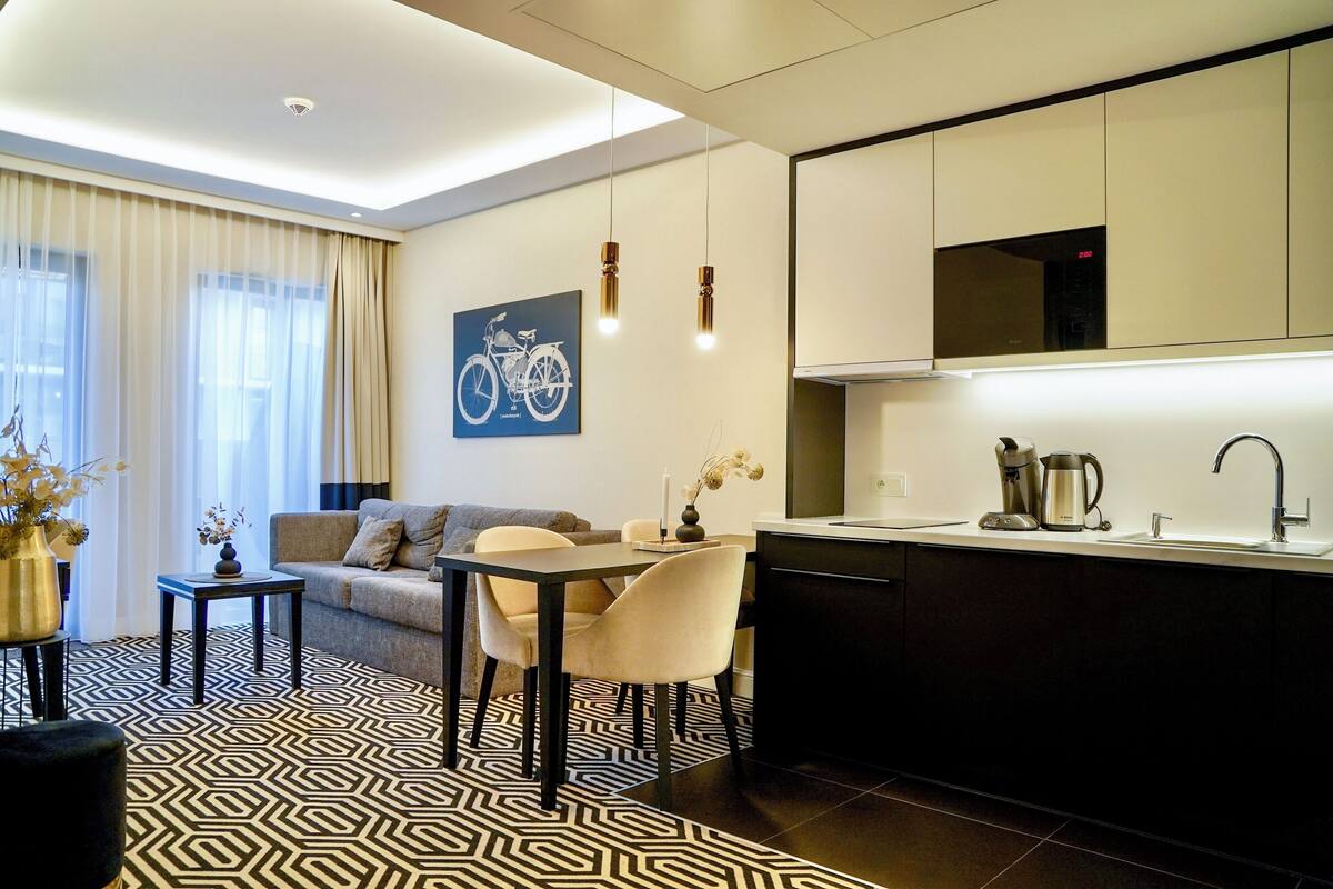 A modern living room with a sofa, dining table, and chairs, a kitchen with a sink and stove, and a patterned rug.