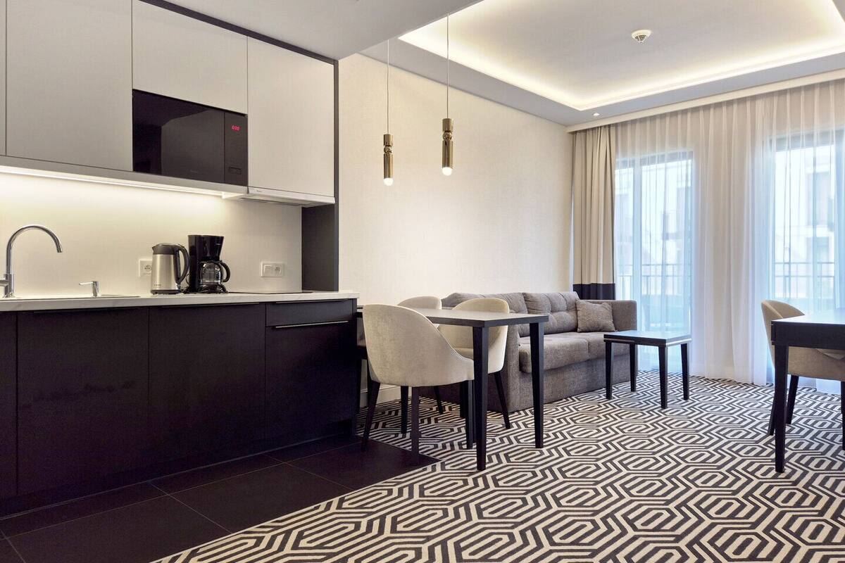 A modern hotel room with a kitchen area, a dining table with chairs, and a patterned floor.