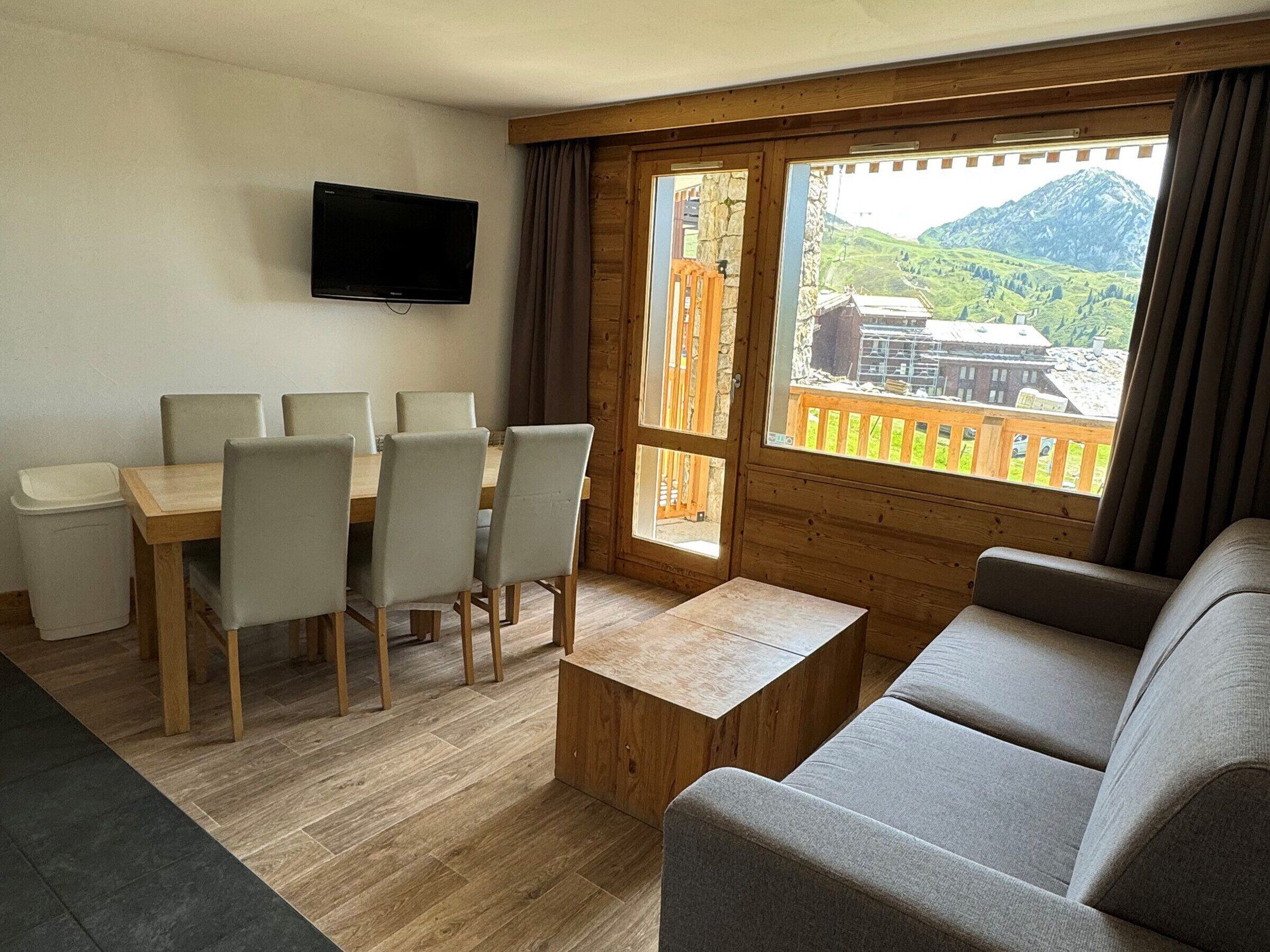 A cozy living room with a wooden dining table, six chairs, a grey sofa, and a TV mounted on the wall.