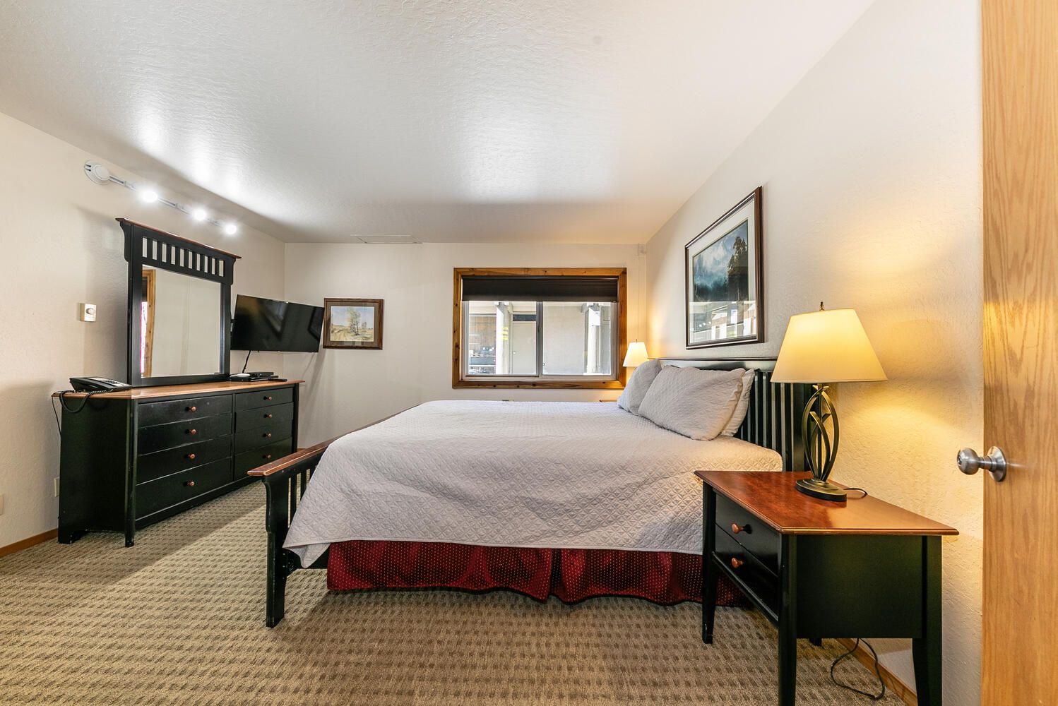 Une chambre d’hôtel comprenant un lit, une commode, un bureau, une lampe, un miroir, une fenêtre et une porte.