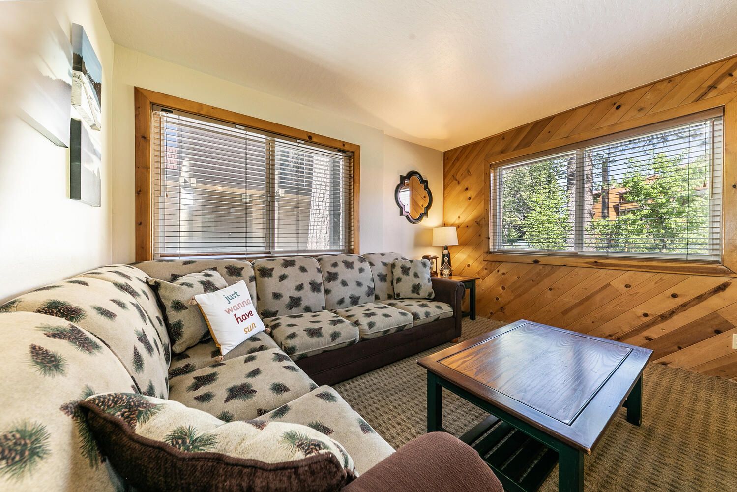 A cozy living room with a sectional sofa, a wooden coffee table, and large windows with blinds.