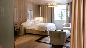 Una habitación de hotel moderna con una cama grande, una zona de estar con una silla y una mesita, un separador de vidrio y un amplio ventanal con cortinas.