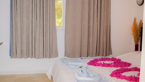 Habitación de hotel con cama, cortinas color beige, una ventana y un arreglo decorativo de flores rosadas en el suelo.
