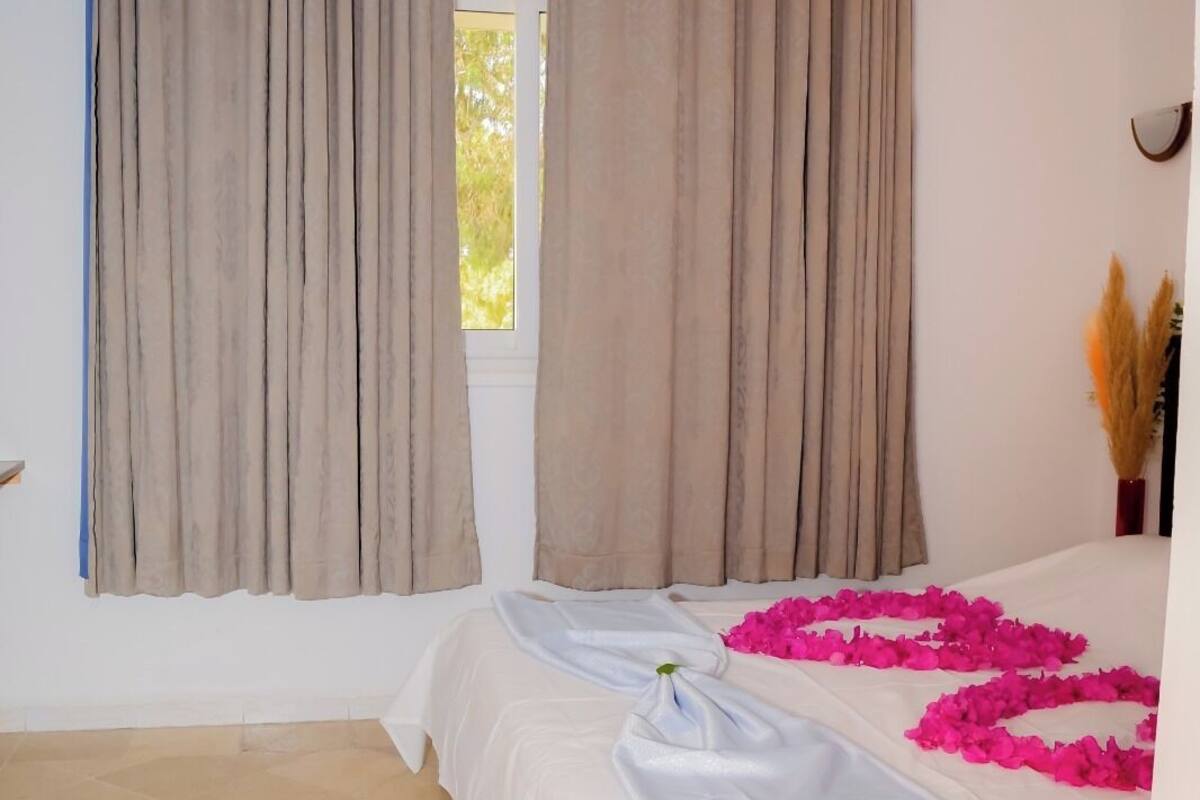 Ein Hotelzimmer mit Bett, beigen Vorhängen, einem Fenster und einer dekorativen Anordnung aus rosa Blumen auf dem Boden.