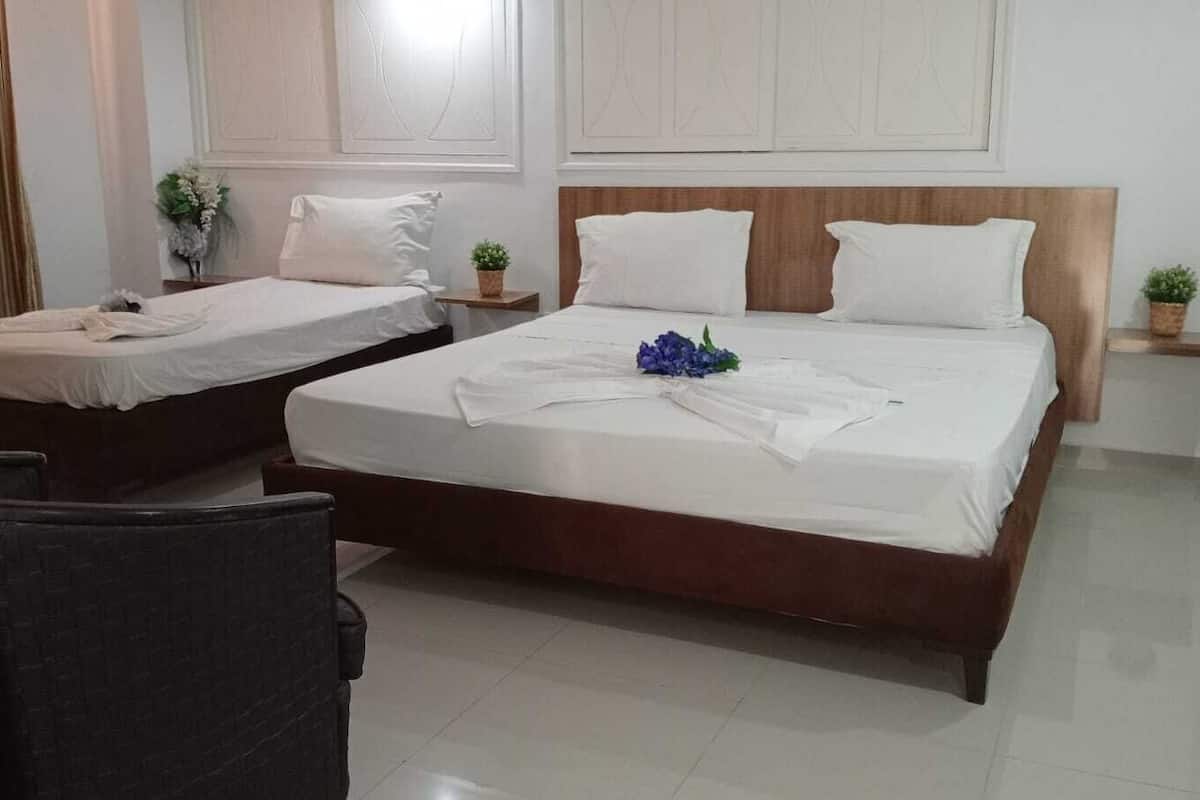 Ein Hotelzimmer mit zwei Einzelbetten, einem großen Bett, einem Holzkopfende, weißen Bettwäsche und einer kleinen Pflanze auf jedem Nachttisch sowie einem dunkel gefärbten Sessel.
