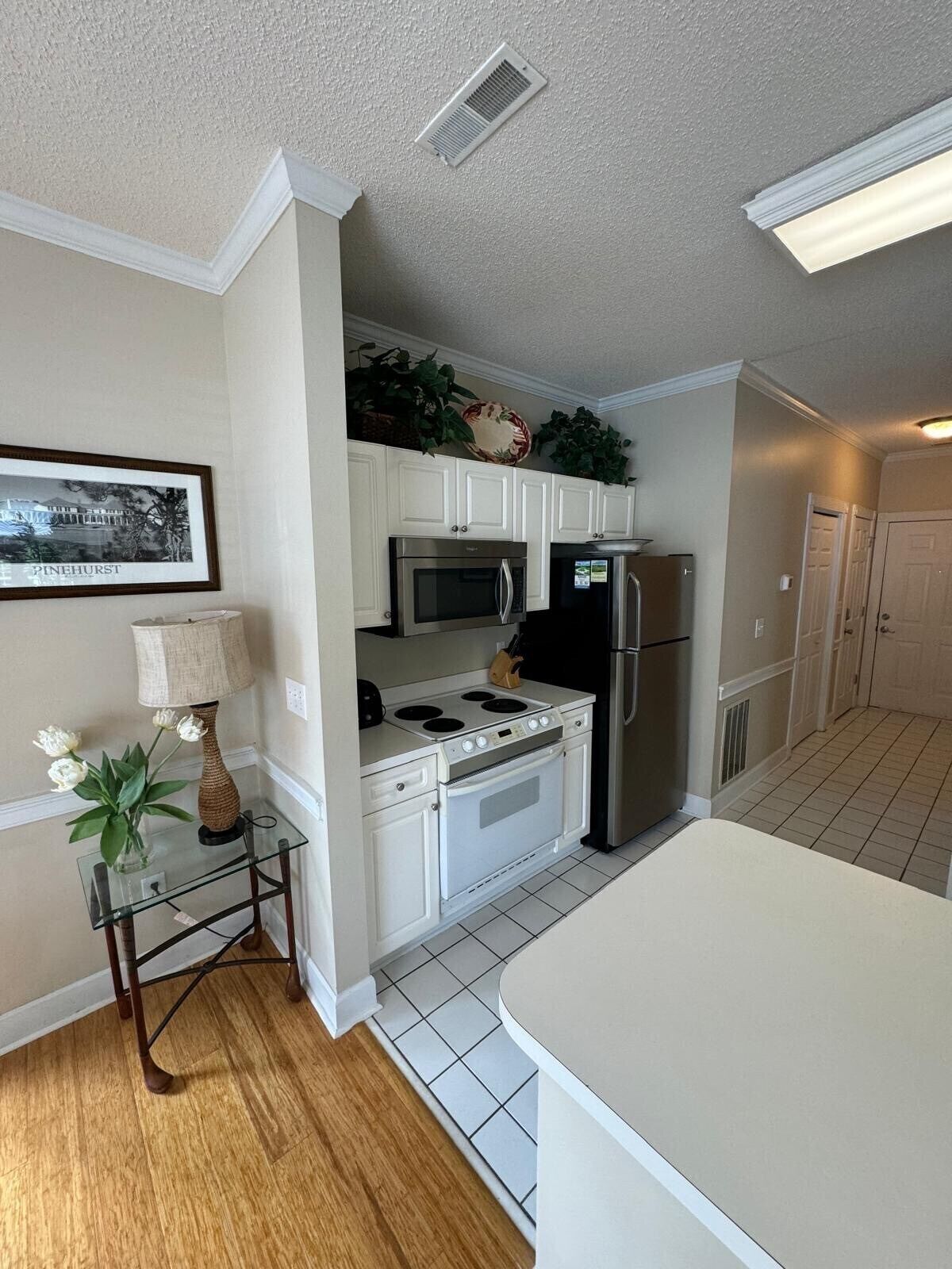 A compact kitchen with white cabinets, a microwave, stove, and refrigerator. There is a small table with a lamp and flowers, and a framed picture on the wall.