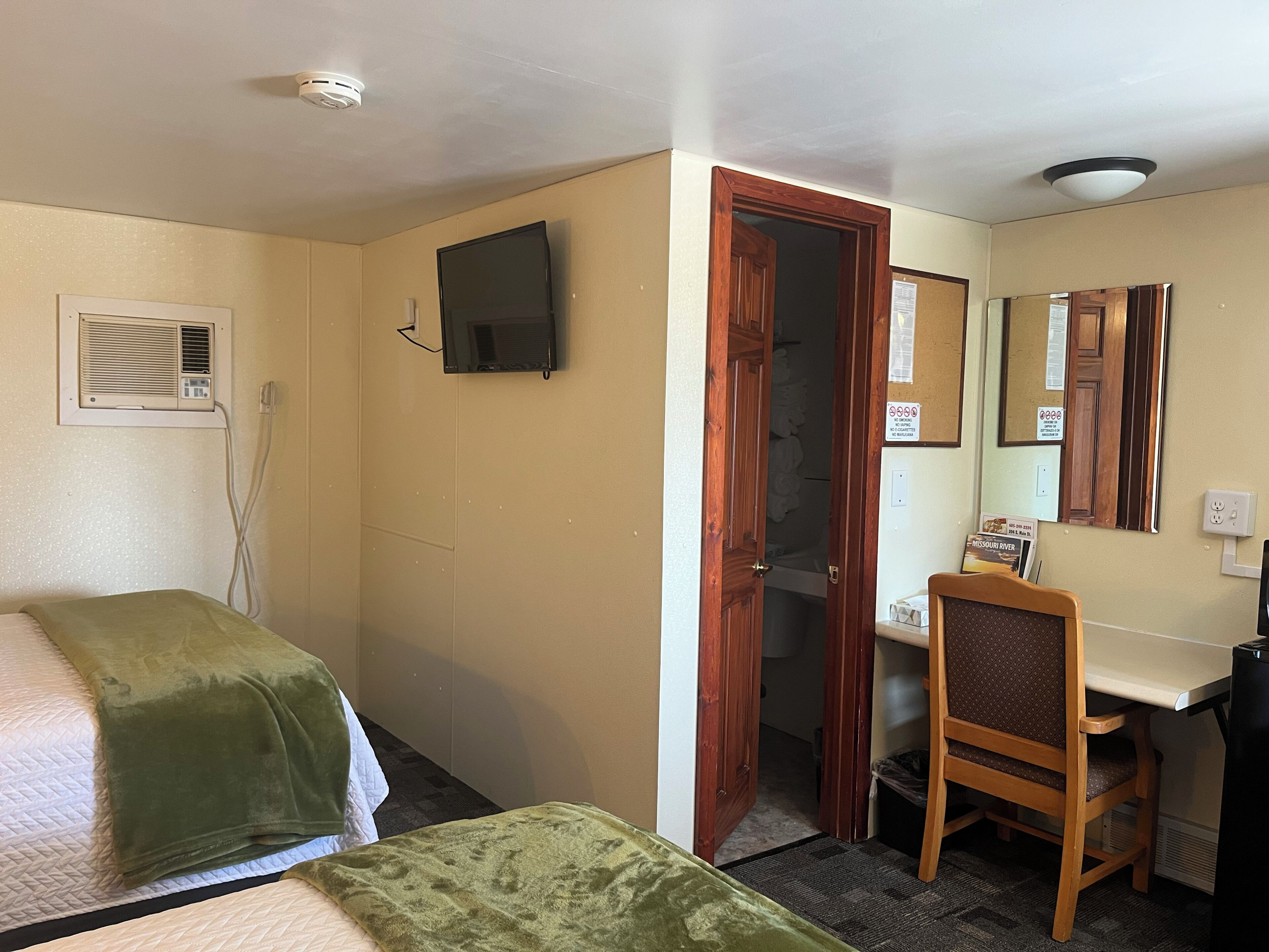 Habitación de hotel con dos camas, un escritorio, una silla, un televisor, un espejo y una puerta que da a otra habitación.