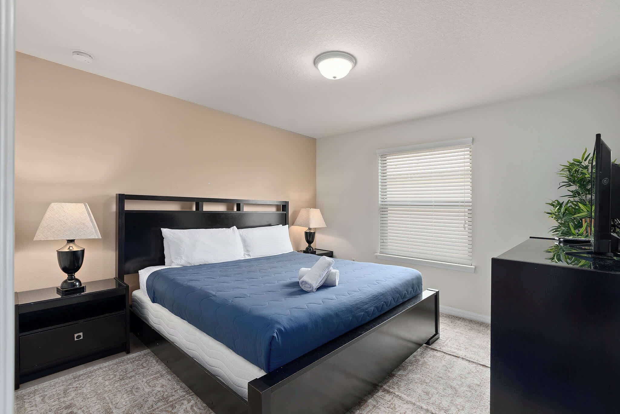A neatly arranged bedroom with a bed, bedside lamps, a nightstand, a dresser, and a window with blinds.