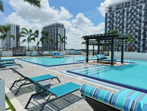 A rooftop swimming pool with lounge chairs and a pergola, surrounded by palm trees and modern buildings.