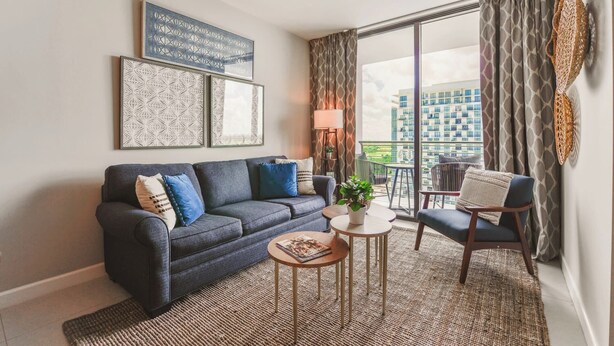 A modern living room with a sofa, a coffee table, and a chair. There are two framed artworks on the wall, a lamp, and a view of the city through large windows. - Prime Downtown Doral Park Condo (Doral)