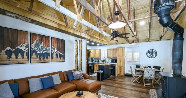 A spacious living area with a brown leather sofa, wooden ceiling beams, a kitchen in the background, and a fireplace. - Yvonnies - Comfy & Renovated Cabin! (Ruidoso)