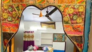 A narrow hallway with a decorative archway leading to a bedroom. The room has a bed with a colorful bedspread, a ceiling fan, and a wall with a framed picture. - Muki Guest House By GRB (Jaisalmer)
