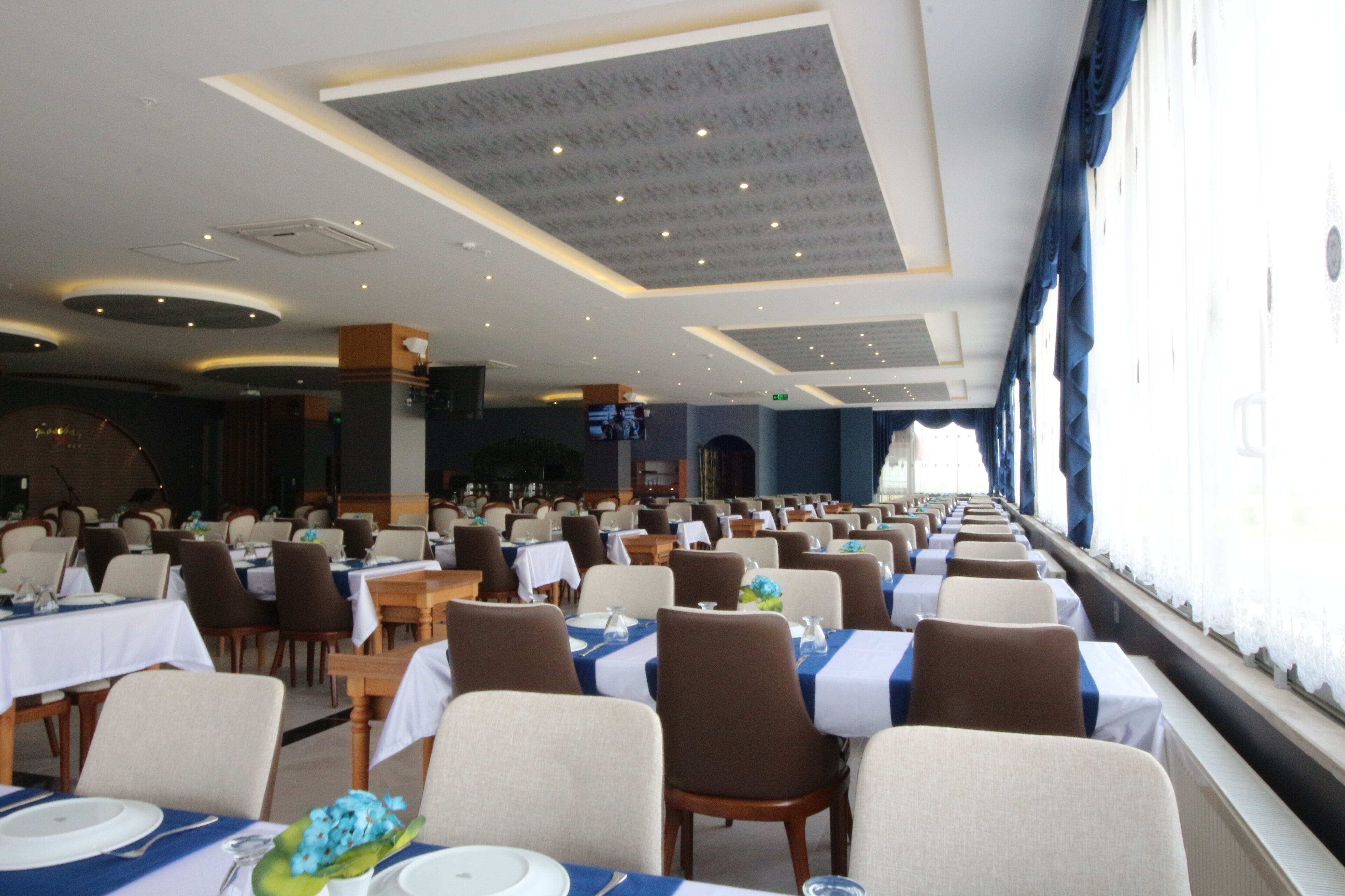 A large dining hall with rows of tables and chairs, a stage in the background, and windows along one wall.