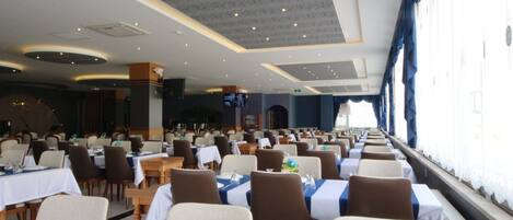 A large dining hall with rows of tables and chairs, a stage in the background, and windows along one wall.