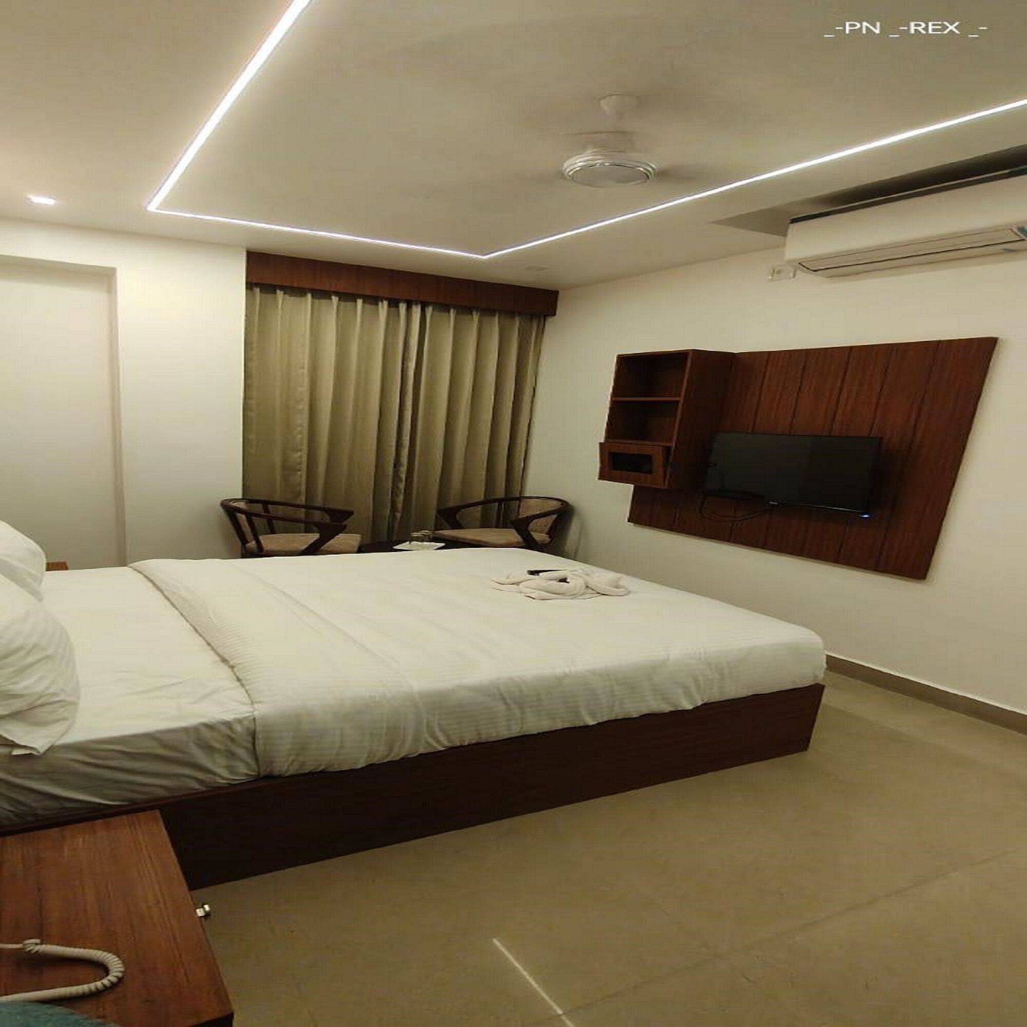 A hotel room with a bed, two chairs, a TV mounted on the wall, and a ceiling light fixture.
