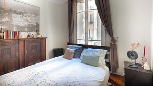 A bedroom with a bed, a dresser, a window with curtains, and a wall-mounted fan. - Opulent 1BD Apartment near Eiffel Tower, Grenelle (Paris)