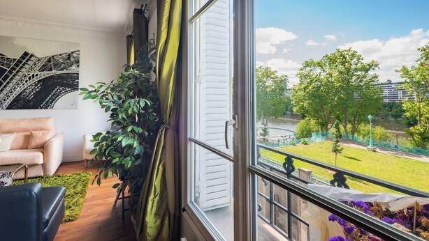 A room with a large window, a sofa, a potted plant, and a view of a park and buildings. - Opulent 1BD Apartment near Eiffel Tower, Grenelle (Paris)