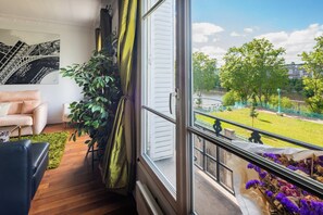 Una stanza con un'ampia finestra, un divano, una pianta in vaso e vista su un parco e degli edifici.