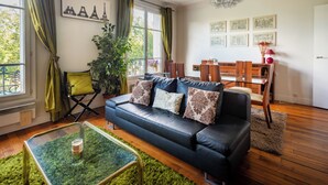 A living room with a black leather sofa, a glass coffee table, a dining table with chairs, and a large window with curtains. - Opulent 1BD Apartment near Eiffel Tower, Grenelle (Paris)