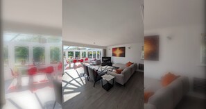 A modern living room with a sofa, coffee table, and TV. There is a dining area with red chairs and a table. Large windows provide natural light and views of greenery. - Thames Riverside Retreat: A Serene 3-Bed Bungalow (Surrey)