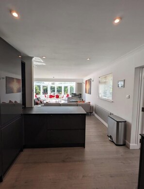 A modern kitchen with a central island, a dining area with red chairs, and a view to an outdoor space. - Thames Riverside Retreat: A Serene 3-Bed Bungalow (Surrey)