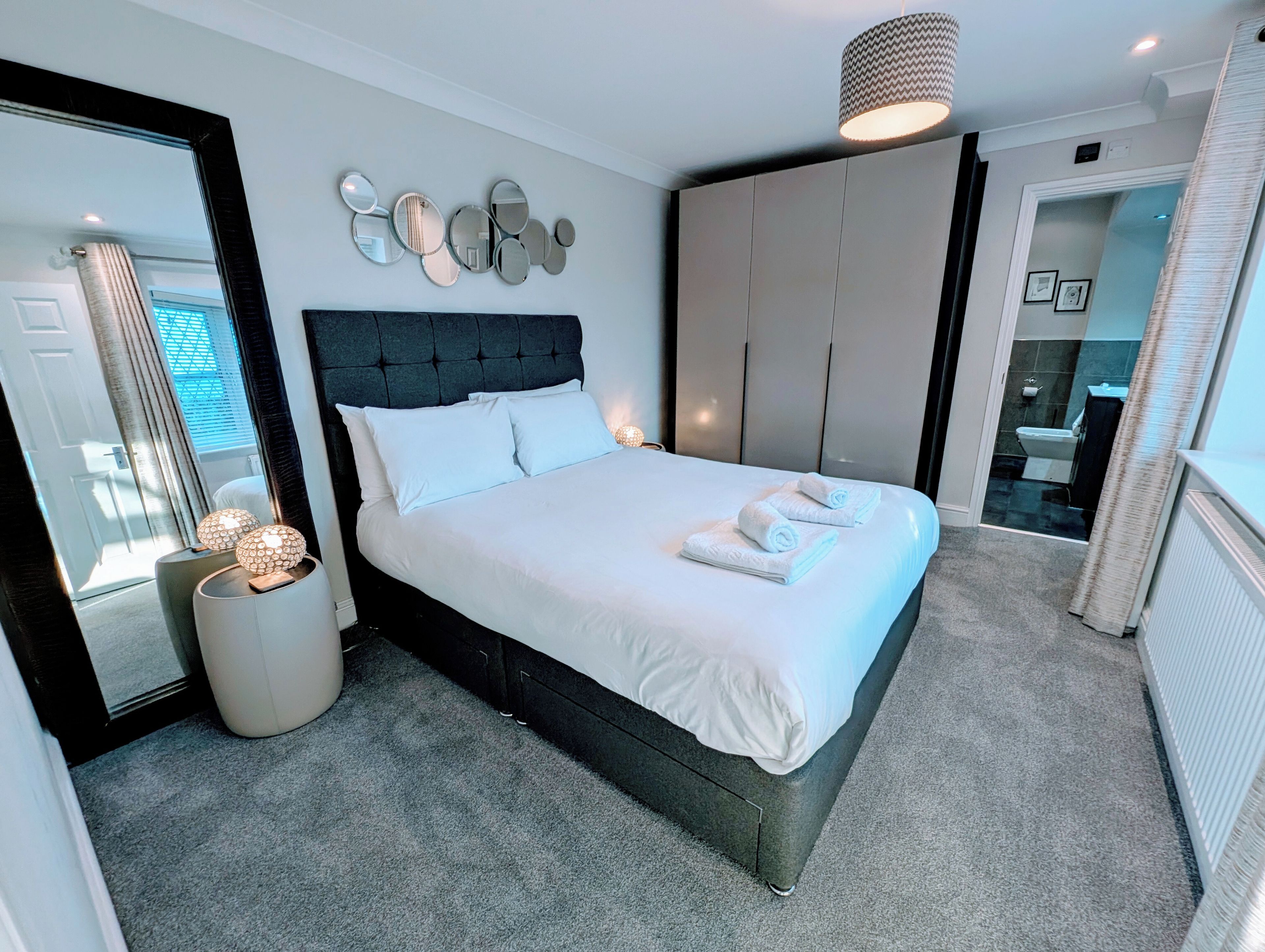 A modern bedroom with a large bed, a headboard, a bedside table with a lamp, a mirror, and a wardrobe.