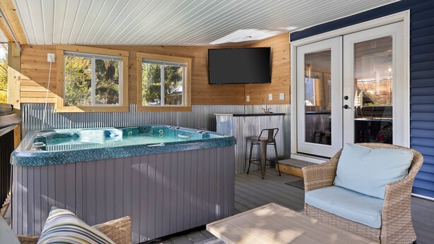 A covered outdoor area with a hot tub, wicker furniture, and a television mounted on the wall. - Walk to Platt River, Game Room, Hot Tub,Indoor Pickleball, Kayaks, Bikes, Arcade (Honor)
