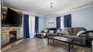 A living room with a stone fireplace, a flat-screen TV mounted on the wall, a grey sofa set, and a coffee table. - Walk to Platt River, Game Room, Hot Tub,Indoor Pickleball, Kayaks, Bikes, Arcade (Honor)