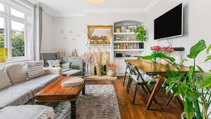 A modern living room with a sofa, coffee table, and dining area. There is a TV mounted on the wall and a bookshelf filled with books and decorative items. - Charming 2BR Family Home with Private Garden (Greater London)