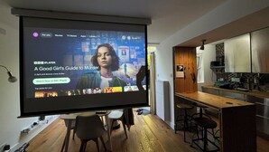 Una sala de estar moderna con una gran pantalla mostrando un programa de televisión, piso de madera, una zona de cocina con taburetes altos y una mesa de comedor.