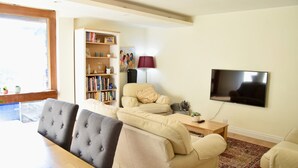 Een woonkamer met een beige bank, een eettafel met stoelen, een boekenkast, een televisie en een lamp.