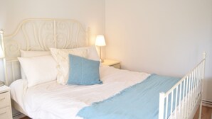 Een net opgemaakt bed met wit- en blauw beddengoed, een wit metalen hoofdbord en een nachtkastje met een lamp.