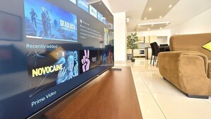 A modern living room with a large flat-screen TV mounted on a wooden wall, a brown sofa, and a dining area in the background. - Private BBQ + outdoor + fun area St. Julians (St Julians)