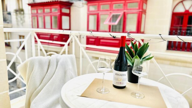 A table with a bottle of wine, two glasses, and a plant on a balcony with white railings and red buildings in the background. - Private BBQ + outdoor + fun area St. Julians (St Julians)