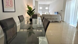 A modern dining area with a glass table and chairs, a potted plant, and a sofa in the background. - Private BBQ + outdoor + fun area St. Julians (St Julians)