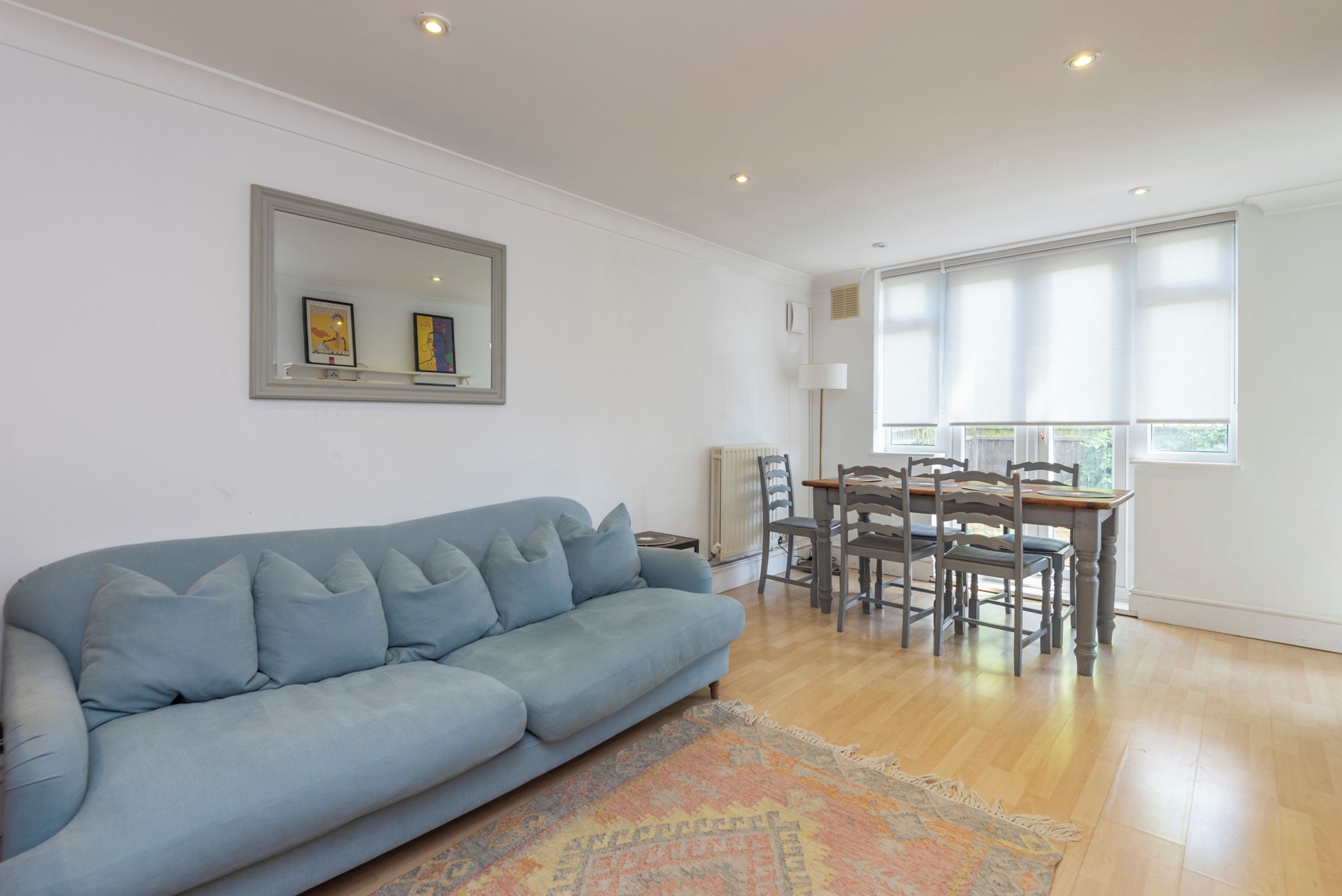 A living room with a light blue sofa, a dining table with chairs, and a large window with blinds.