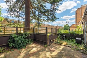 Un patio trasero cercado con un árbol grande, una parrilla y un edificio de ladrillo al fondo.