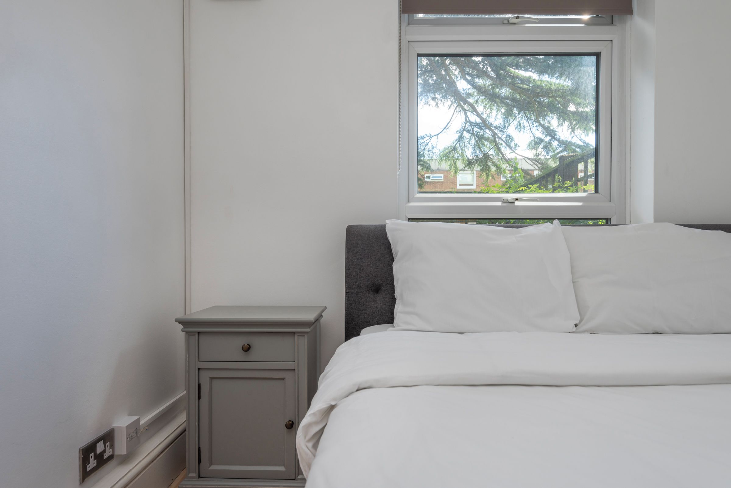 A bedroom with a bed, a nightstand, a window with a view of trees, and a wall socket.