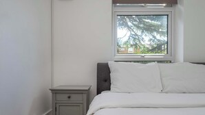 Ein Schlafzimmer mit einem Bett, einem Nachttisch, einem Fenster mit Blick auf Bäume und einer Wandsteckdose.