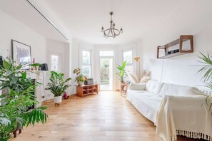 A bright room with a wooden floor, white walls, a white sofa, and various plants. - Modern Farmhouse-Style 1BD Flat - Wood Green! (Greater London)