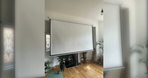 Una habitación con una gran pantalla blanca, suelo de madera, una pequeña mesa y una planta en maceta.
