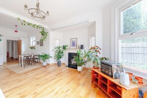 Uma sala de estar ampla com lareira, piso de madeira e diversos vasos de plantas.