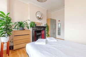 A bedroom with a bed, a dresser, a mirror, a fireplace, and several potted plants. - Modern Farmhouse-Style 1BD Flat - Wood Green! (Greater London)