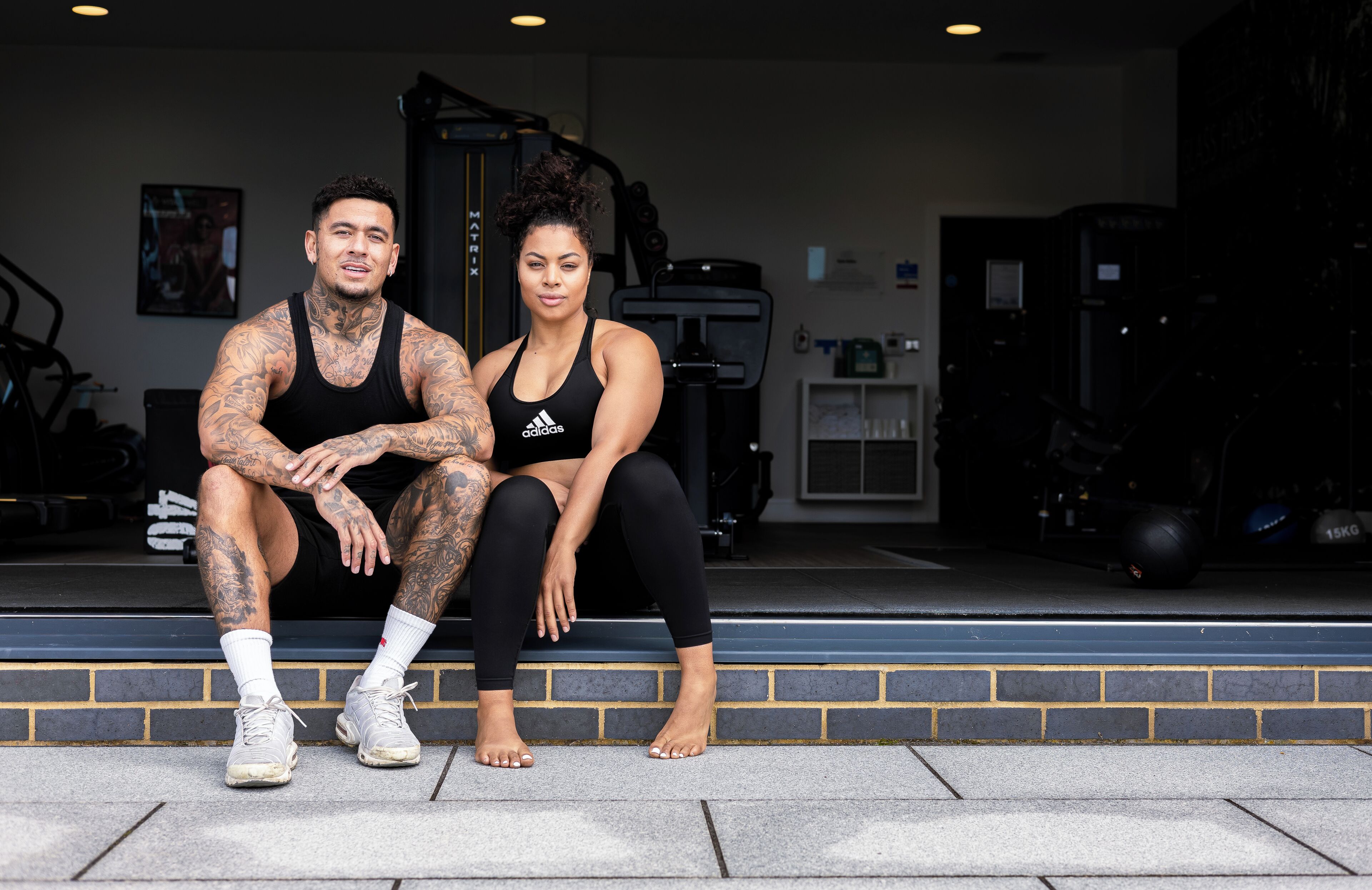 Ein Mann und eine Frau sitzen vor einem Fitnessstudio auf Stufen, im Hintergrund Trainingsgeräte.